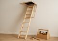 A wooden loft ladder extending from an attic entrance with a toolbox beside it.